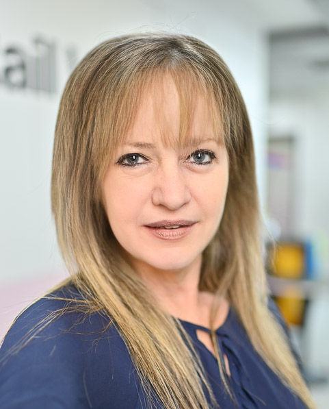 A woman with shoulder-length blonde hair and a blue top is looking directly at the camera with a slight smile. The background is blurred.
