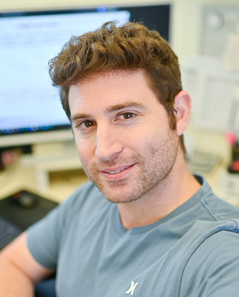 A man with wavy brown hair and a beard is sitting at a desk, smiling at the camera. A computer monitor and papers are visible in the background.