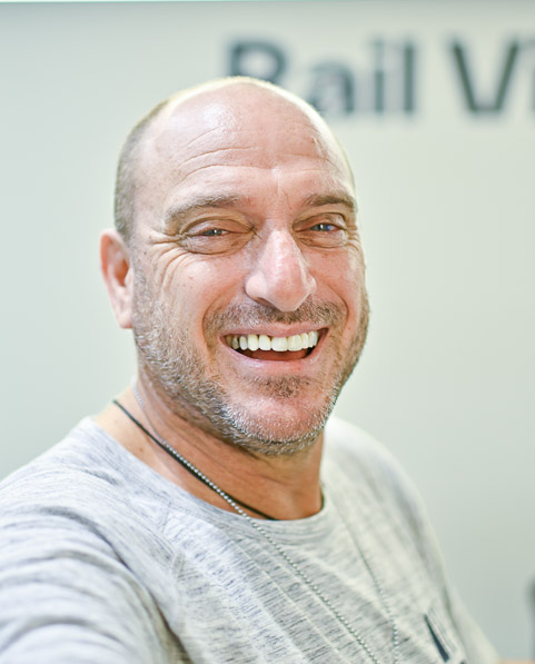 A bald man with a gray beard and mustache smiles at the camera, wearing a gray shirt and a necklace.