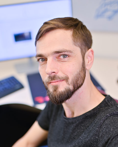 A man with a beard and short hair taking a selfie in an office setting with a computer monitor and keyboard in the background.