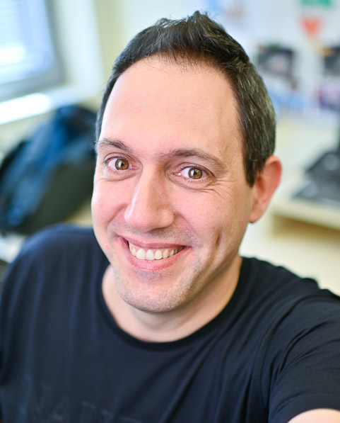 A man with short dark hair wearing a black t-shirt smiles at the camera in an office setting.