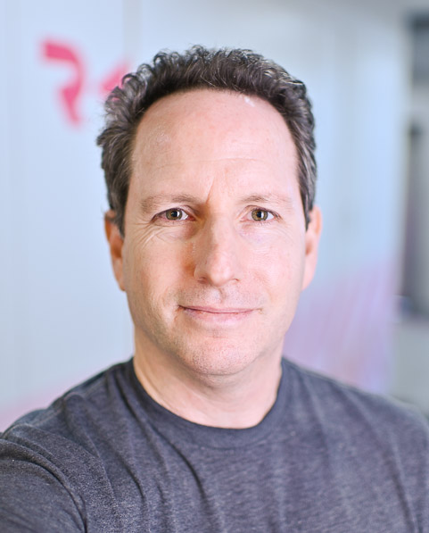 A person with short, dark hair, wearing a grey t-shirt, standing indoors with a blurred background.