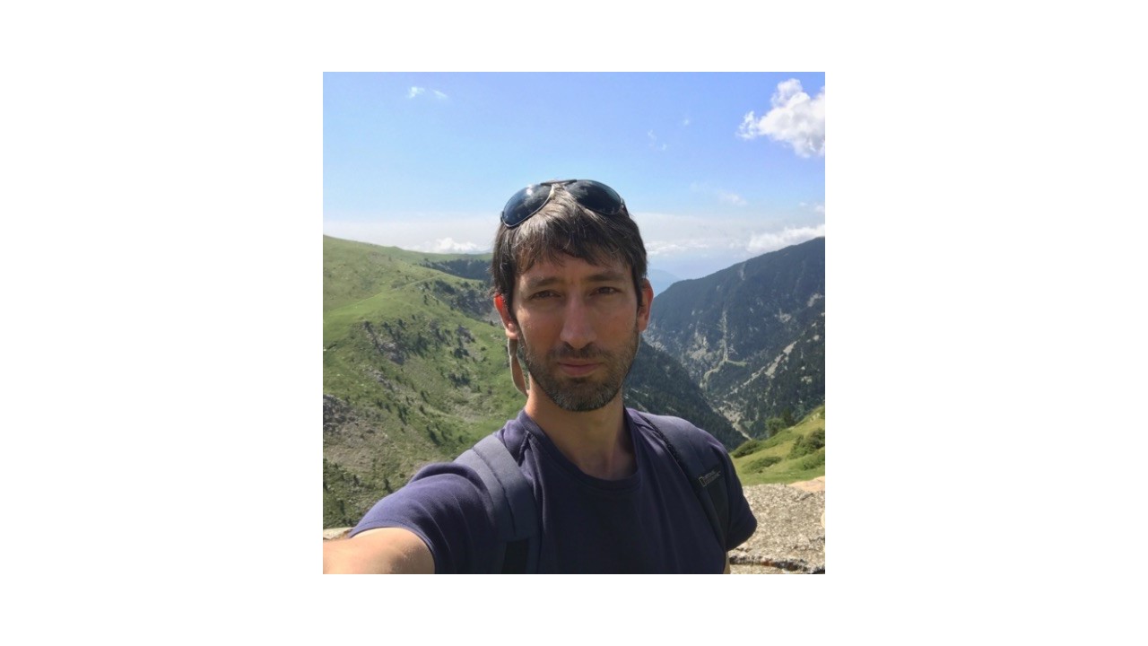 A man with dark hair and sunglasses on his head takes a selfie on a hiking trail with mountains and a clear blue sky in the background.