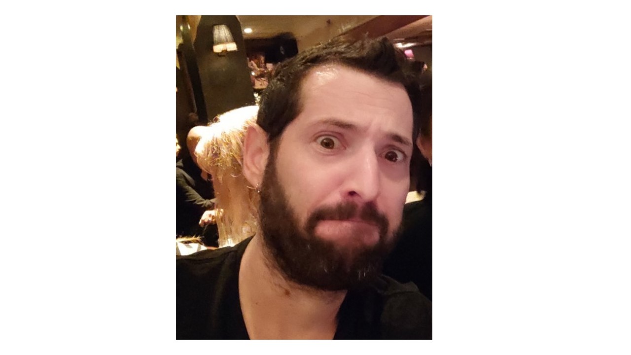 A man with a beard and mustache is posing with a concerned expression in a dimly lit indoor setting.